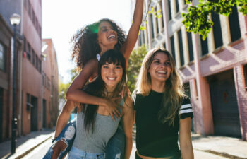 Three young women outdoors on a city street, one giving another a piggyback ride while all three smile and enjoy the sunny day.