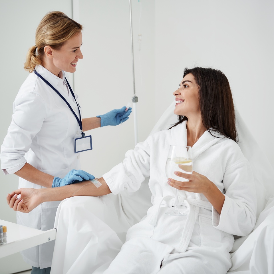 Portrait of attractive woman in white bathrobe sitting in armchair and holding glass of lemon water while physician in sterile gloves checking IV infusion