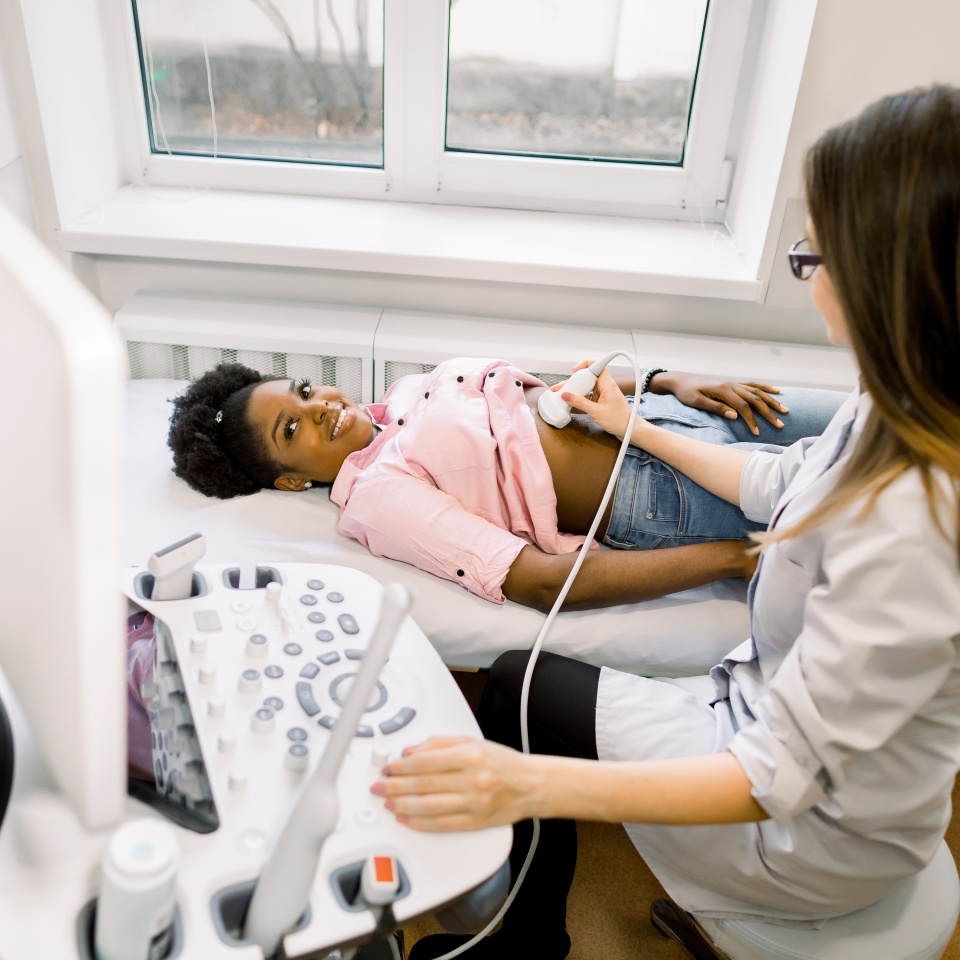 Ultrasound diagnostic of stomach on abdominal to young African woman. Doctor runs ultrasound sensor over patient's tummy and working on scanner panel. Diagnosis of internal organs.