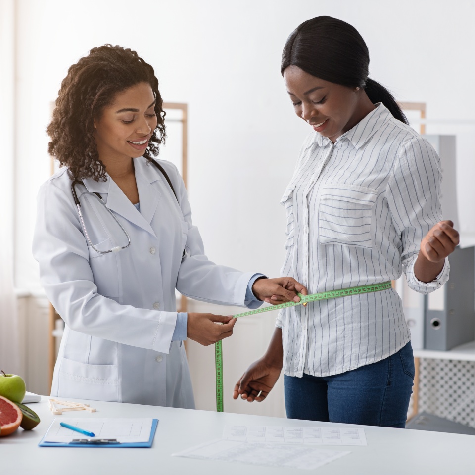 Young black lady visiting modern dietologist, taking body measurements, clinic interior