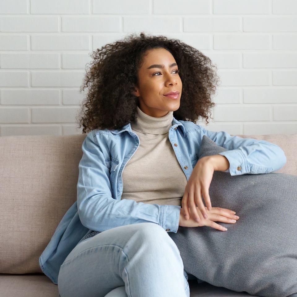 Happy satisfied relaxed young african woman lounge on comfortable modern furniture sofa. Smiling black girl sitting on couch in cozy living room looking away, dreaming, feel no stress at home concept.