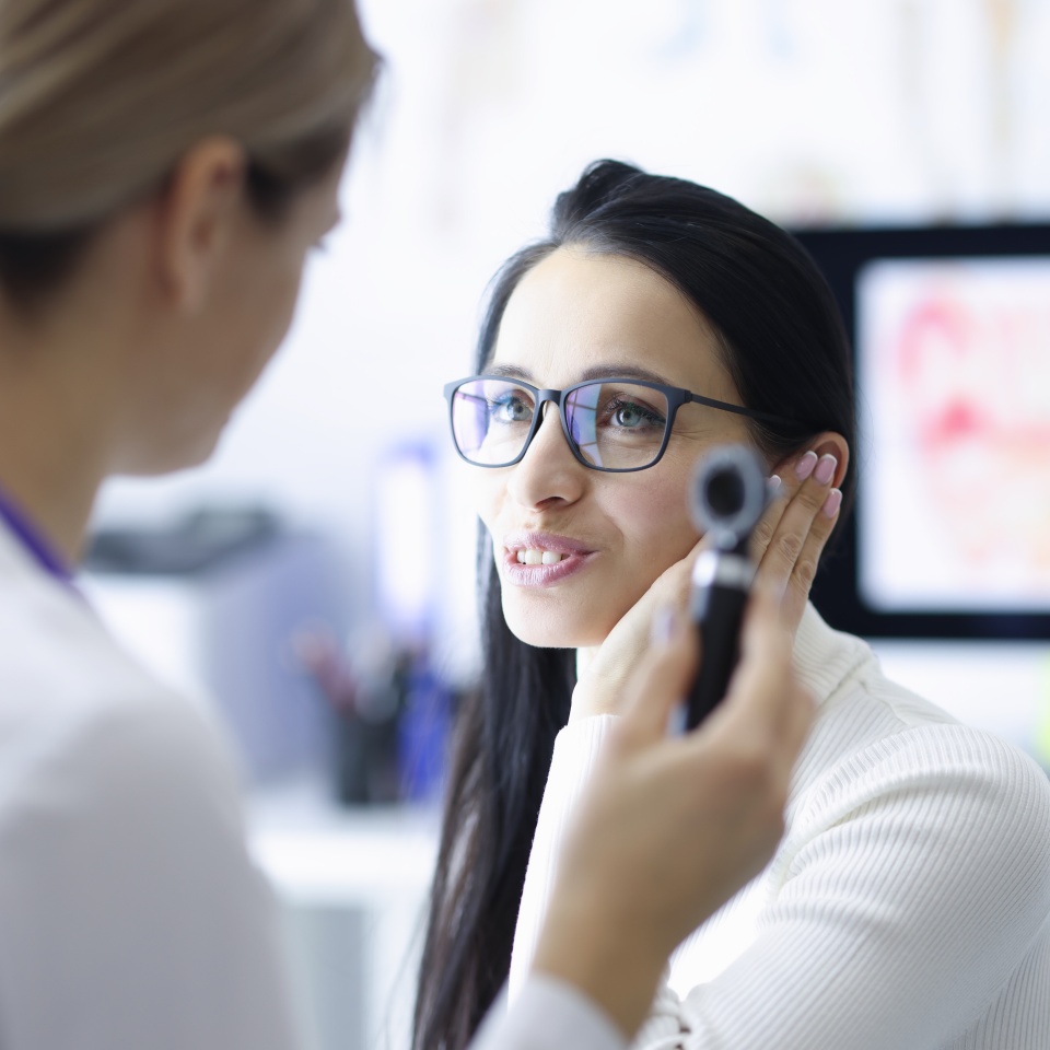 Woman patient holding ear near doctor with otoscope in hand in clinic. Diagnosis and treatment of hearing loss concept