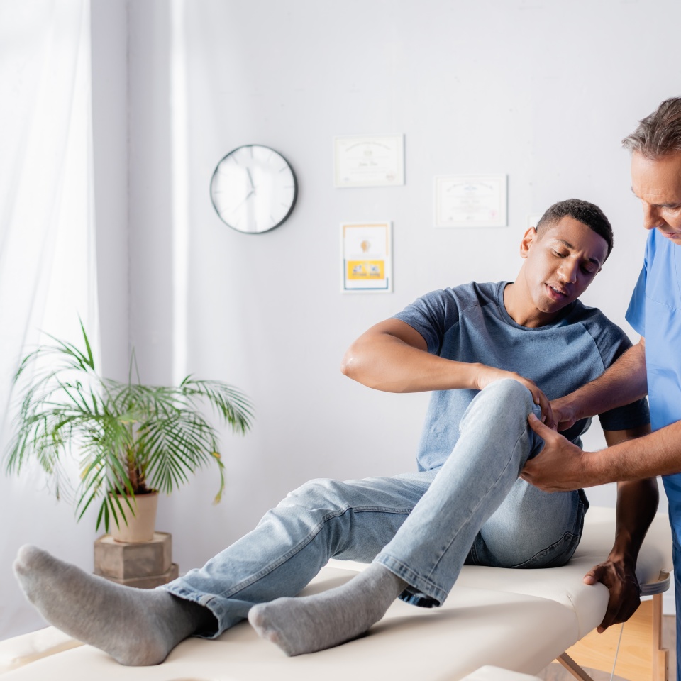 mature chiropractor touching injured knee of african american patient