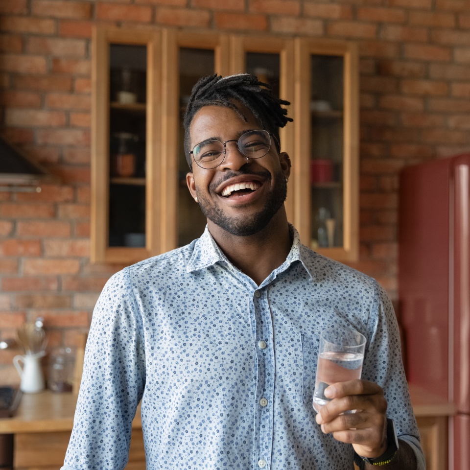 Head shot portrait overjoyed African American man holding glass of fresh pure mineral water, standing in kitchen, looking at camera, healthy lifestyle and good daily habit concept, body and skin care