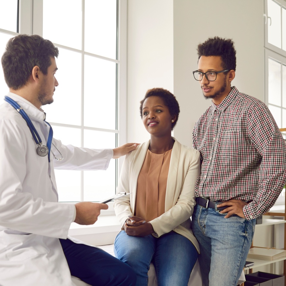 Family visiting doctor for health check up. Professional gynecologist, OB GYN obstetrician, general practitioner or IVF pregnancy specialist talking to young black couple, reassuring and giving hope