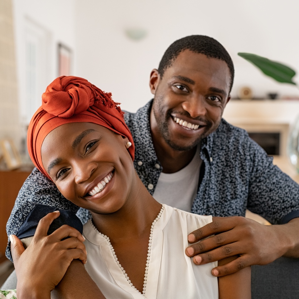 Portrait of mid adult happy african american couple hugging at home while sitting on couch. Portrait of mature man with his woman wearing traditional turban looking at camera. Black mid adult couple.