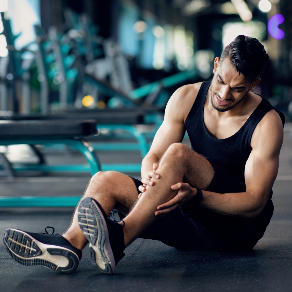 Sport Injury. Upset Arab Male Athlete Suffering From Knee Trauma While Training At Gym, Middle Eastern Man Sitting On Floor And Massaging Aching Area, Sporty Guy Frowning With Pain, Copy Space