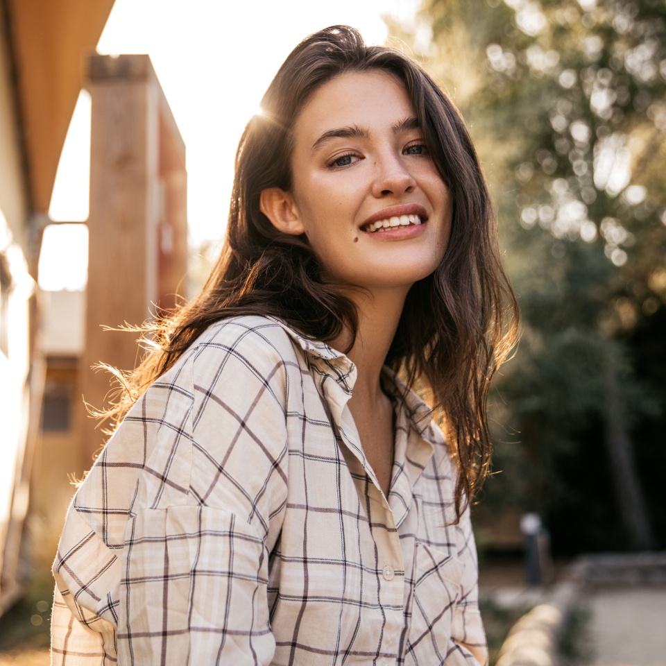 Pretty young caucasian woman enjoying spending time outdoors in sunny weather. Brunette looks at camera, wears shirt. Relaxation concept