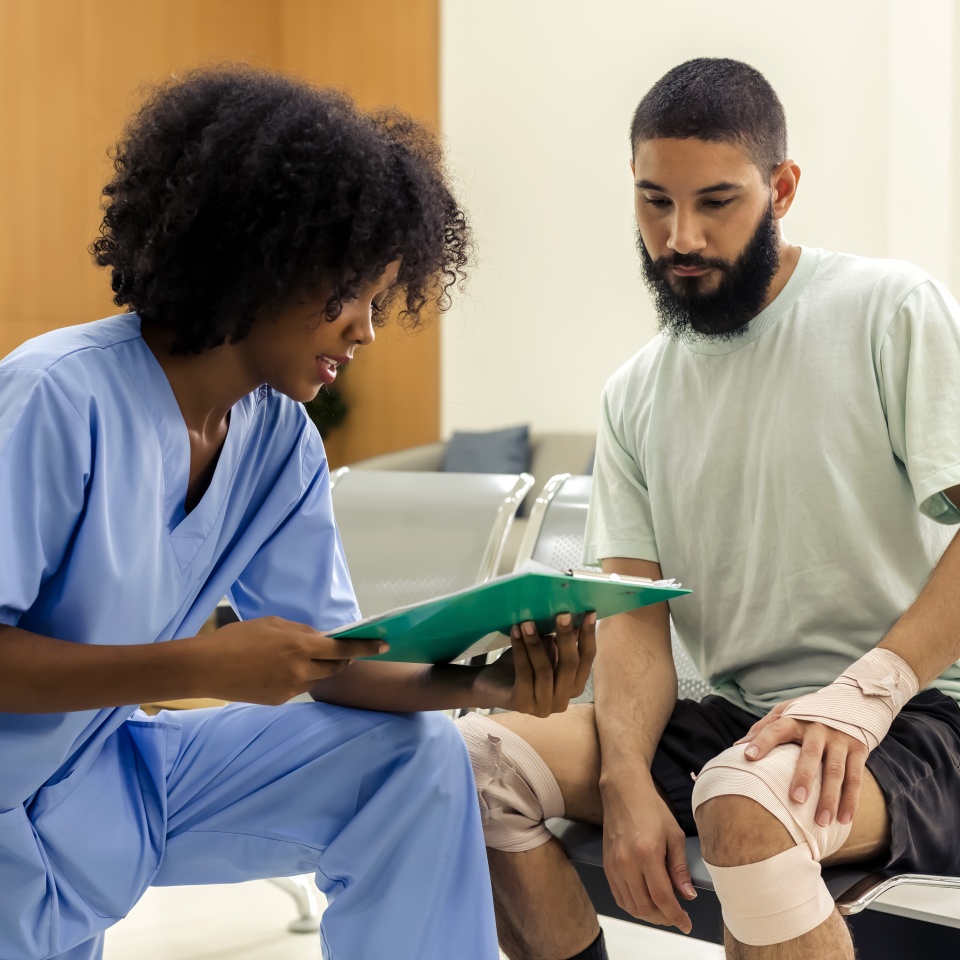 African black nurse woman asking injured informations from young Caucasian patient man in hospital, female afro hair nurse in blue hospital uniform checking beard man patient about injured wound