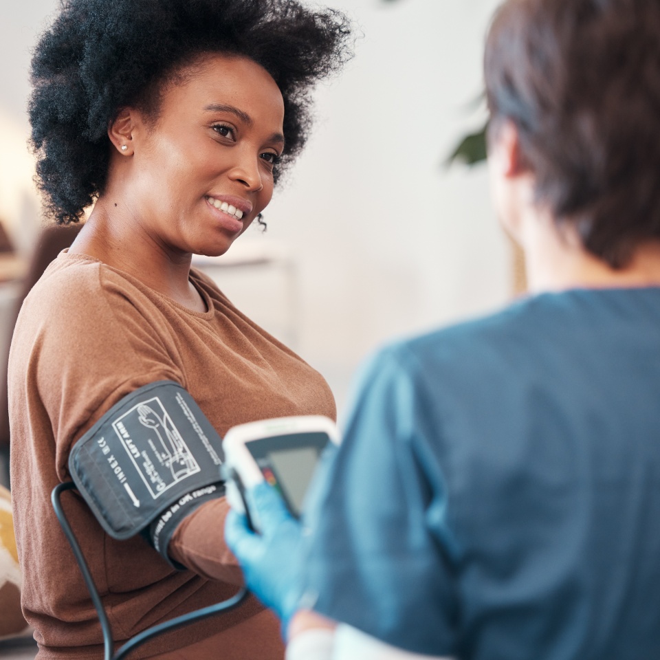 Black woman, healthcare and blood pressure checkup with caregiver monitoring or checking pulse at home. Happy African American female patient or visit from medical nurse for health and wellness