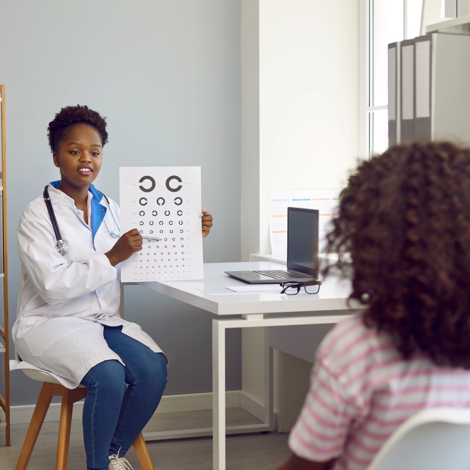 Child is at ophthalmologist's appointment. Female optometrist examines vision of child girl patient using vision chart in hospital office. African American woman doing medical examination of child.