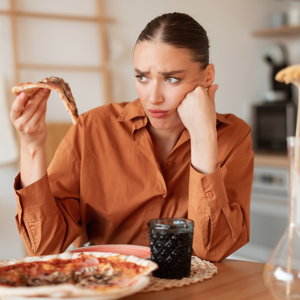 Upset woman looking at delicious piece of high-calorie pizza, holding slice in hand. Diet and digestive problems as well as spoiled and tasteless food