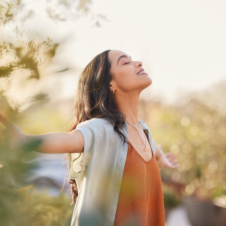 Young latin woman with arms outstretched breathing in fresh air during sunrise at the balcony. Girl enjoying nature while meditating during morning with open arms and closed eyes. Mindful woman relax.