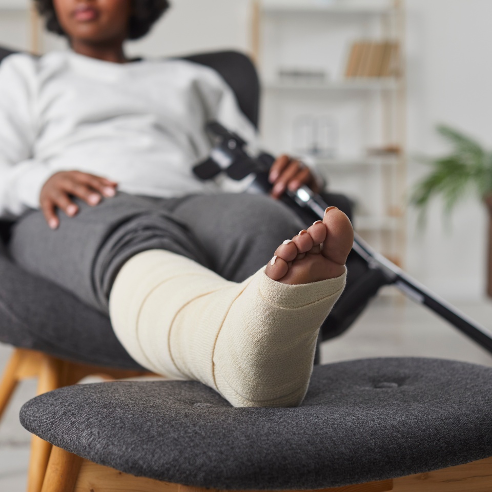 African american woman is sitting at home in armchair with broken bandaged leg lying on stool and holding crutches in her hands. Recovery time after physical injury of the foot bone. Selective focus.
