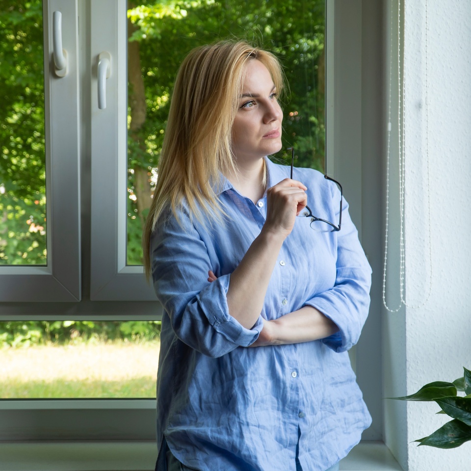 Close-up positive portrait Beautiful middle-aged blond woman with glasses stands near the window at home, looks at the camera and smiles. Copy space