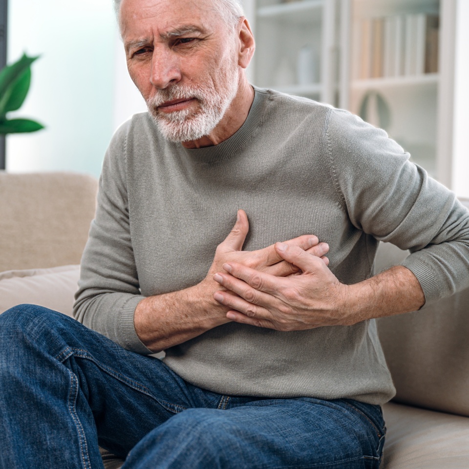 mature man feeling unwell, cardiac pain in chest, sitting on couch at home and need medical help. senior male has heart attack symptom. healthcare and medicare concept
