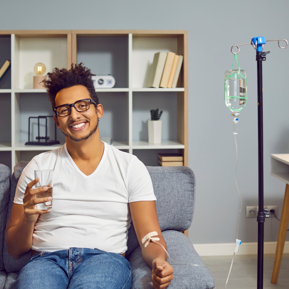 Young happy african american man sitting on the couch in the clinic holding a glass of water while receiving IV drip infusion and vitamin therapy in his blood. Person receiving injection therapy.