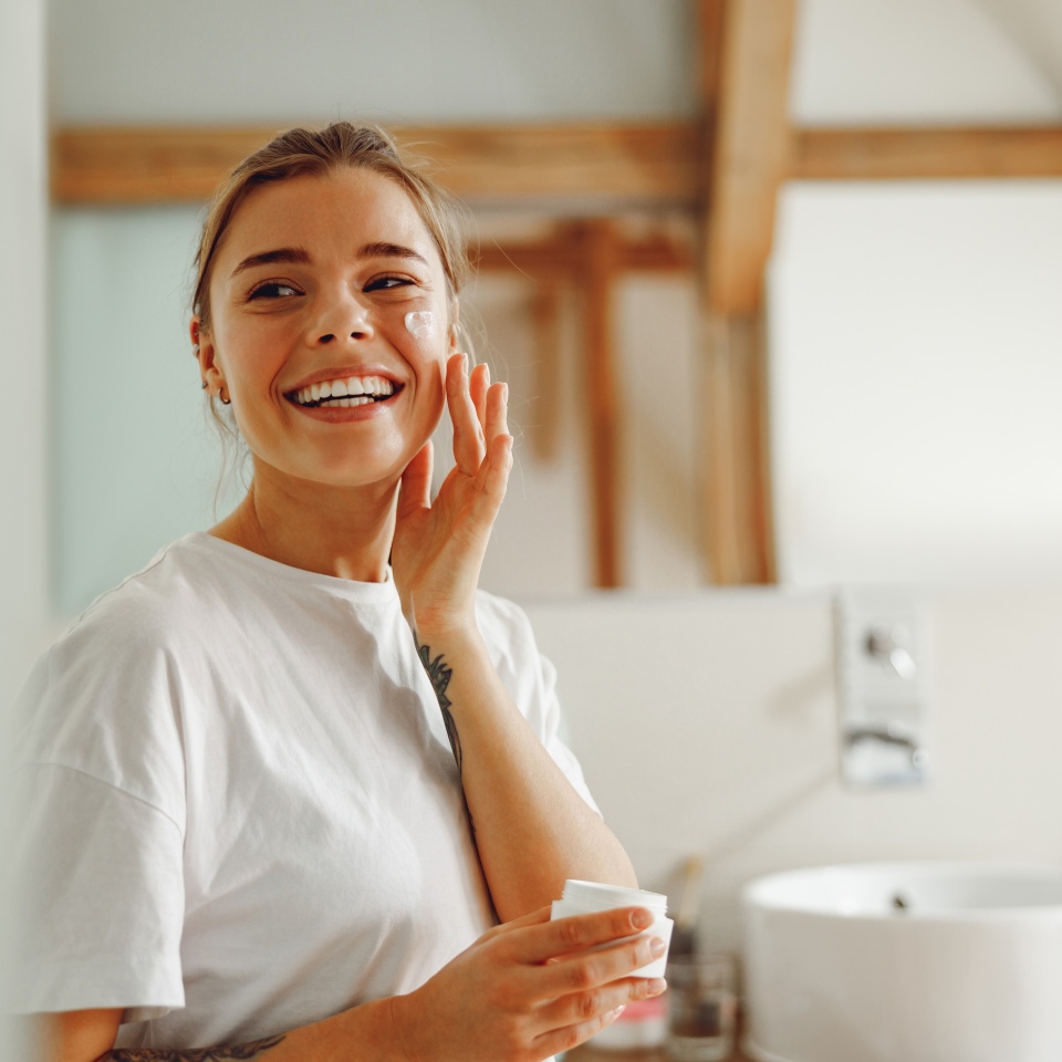 Beautiful young woman taking care of skin by applying moisturizer cream in bathroom