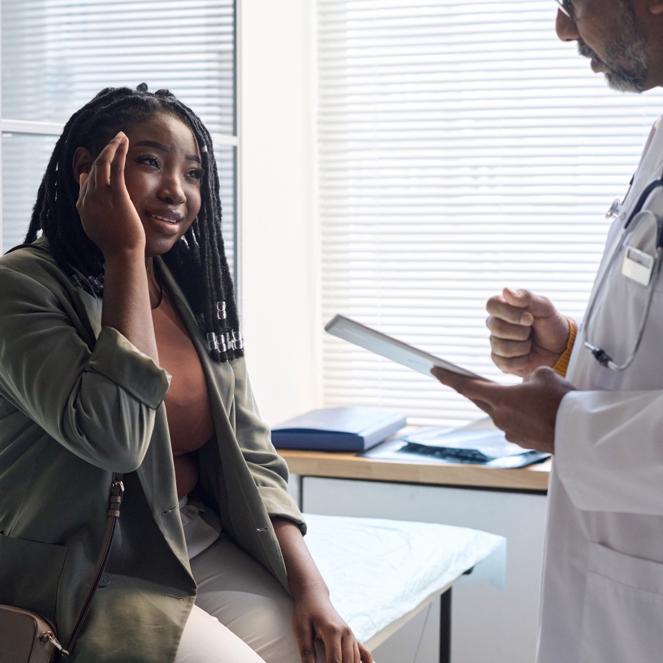 Portrait of African American woman suffering from headache explaining symptoms to doctor during consultation in clinic copy space