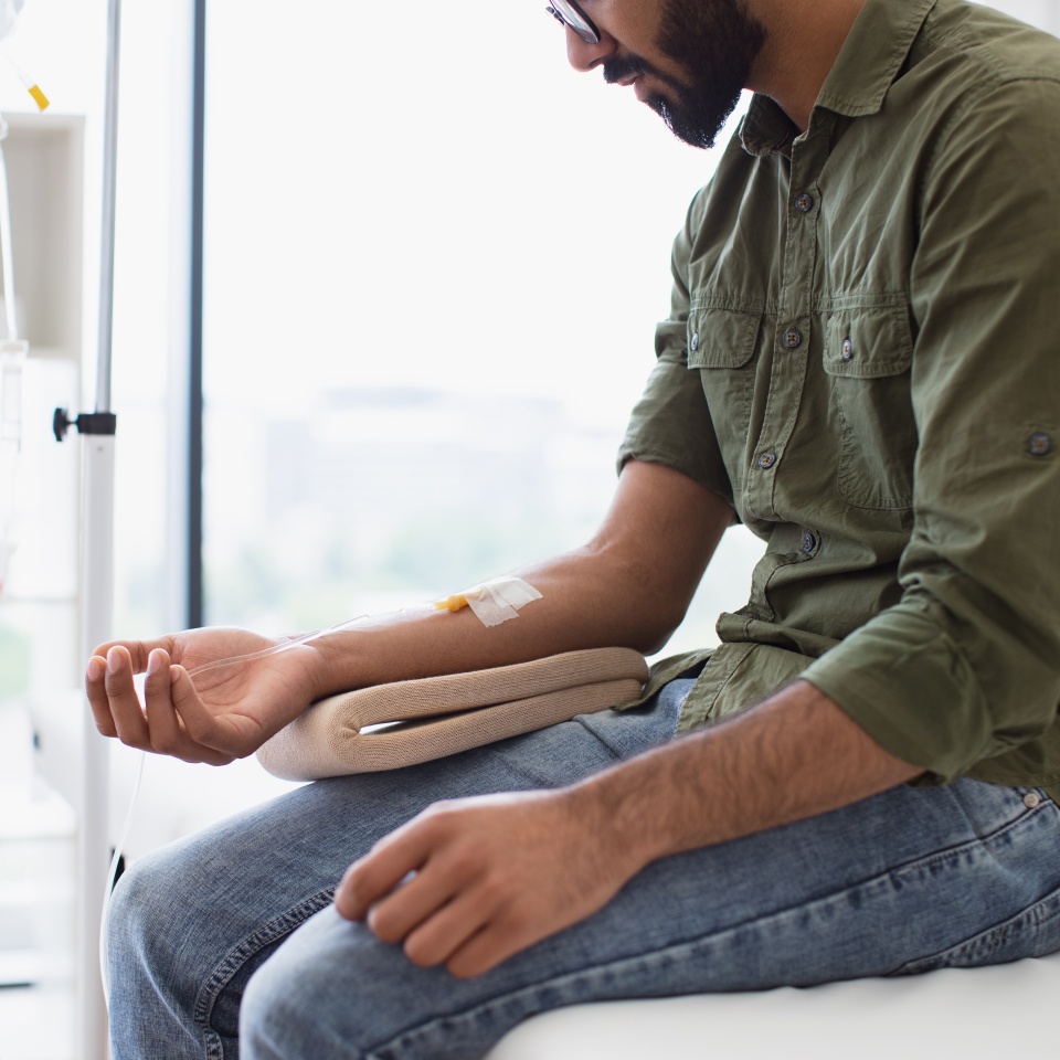 Young bearded man taking dropper with solution for chemotherapy sitting on exam couch. Tired sick male in casual wear drips medicine intravenously while sitting on modern light ward.