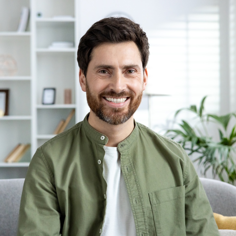 Portrait of a smiling man sitting on a couch in a cozy living room, with shelves and plants in the background.