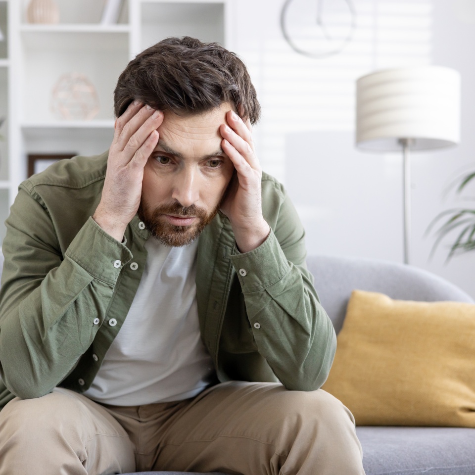 Stressed man sitting on sofa holding his head, feeling overwhelmed and anxious. Man experiencing mental health issues, worry, frustration, and emotional stress. Concept of anxiety and mental health