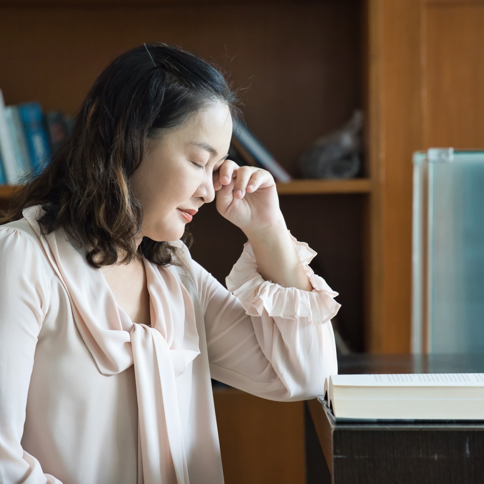 Stressed Asian Middle-Aged Woman Reading a Difficult Book in a Quiet Library, having dry eye, eye soreness