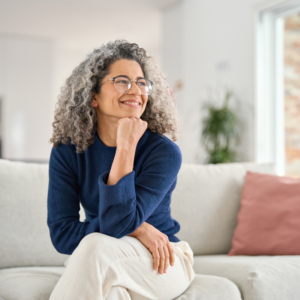 Happy relaxed 50 years old woman sitting on sofa at home looking away. Smiling middle aged lady posing in living room at home. Mature older female with curly gray hair relaxing on couch enjoying chill