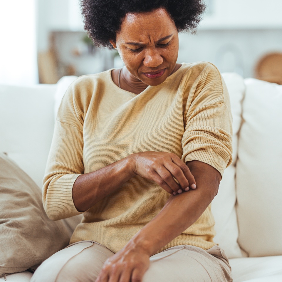A woman sitting on a couch scratching her arm, indicating skin irritation or itchiness. The setting is a comfortable home environment, highlighting discomfort and potential skin issues.