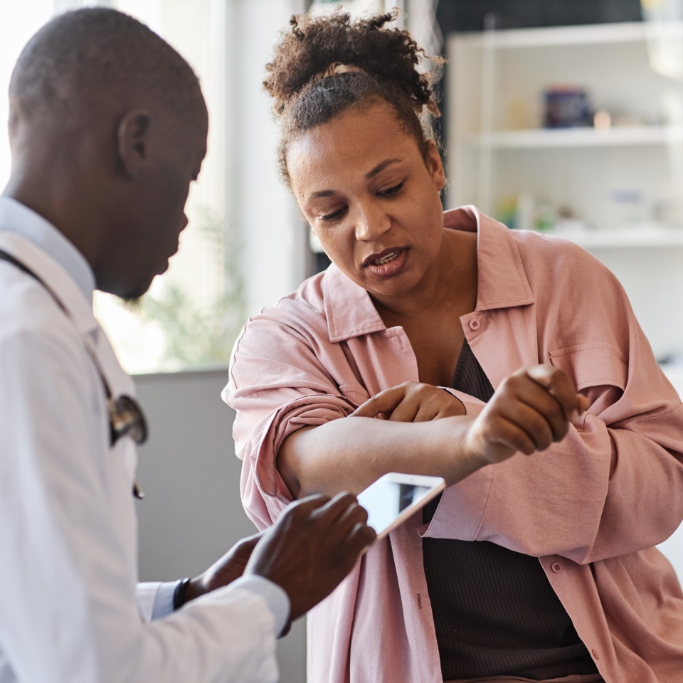 Adult African American woman suffering from psoriasis visiting dermatologist in clinic and pointing on rash spots on arms copy space