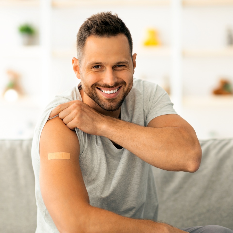 Global immunization benefits. Happy young vaccinated man showing shoulder with medical plaster after vaccine injection and smiling widely to camera, sitting on sofa at home