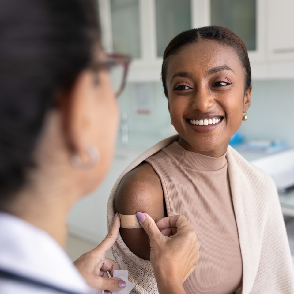 Cheerful young African patient woman visiting doctor for vaccination, talking to physician, nurse sticking plaster on shoulder after injection, smiling, getting vaccine for protection from flu virus