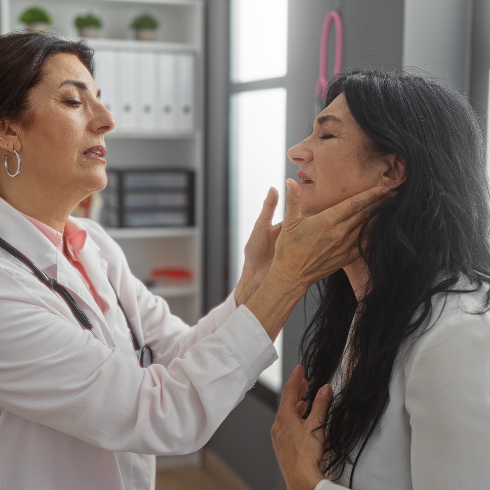 Mature female doctor examines woman's throat in a modern clinic interior, showcasing medical professionalism and patient care dynamics in a well-lit workplace.