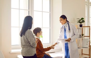 Portrait of a friendly smiling female doctor pediatrician shaking hand with a little boy patient standing with his mother and listening the recommendations after medical examination.