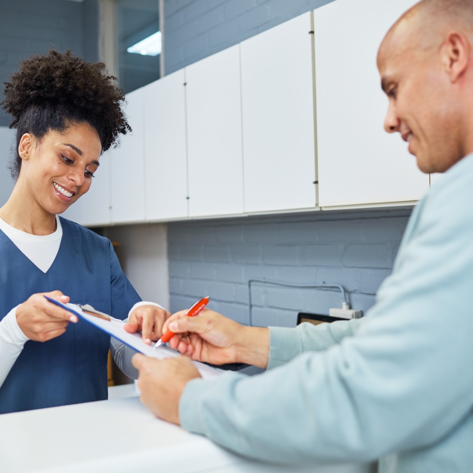 A cheerful medical professional dressed in scrubs helps a patient complete paperwork at a clinic's reception area, creating a welcoming atmosphere that highlights attentive customer service and professionalism in healthcare environments.