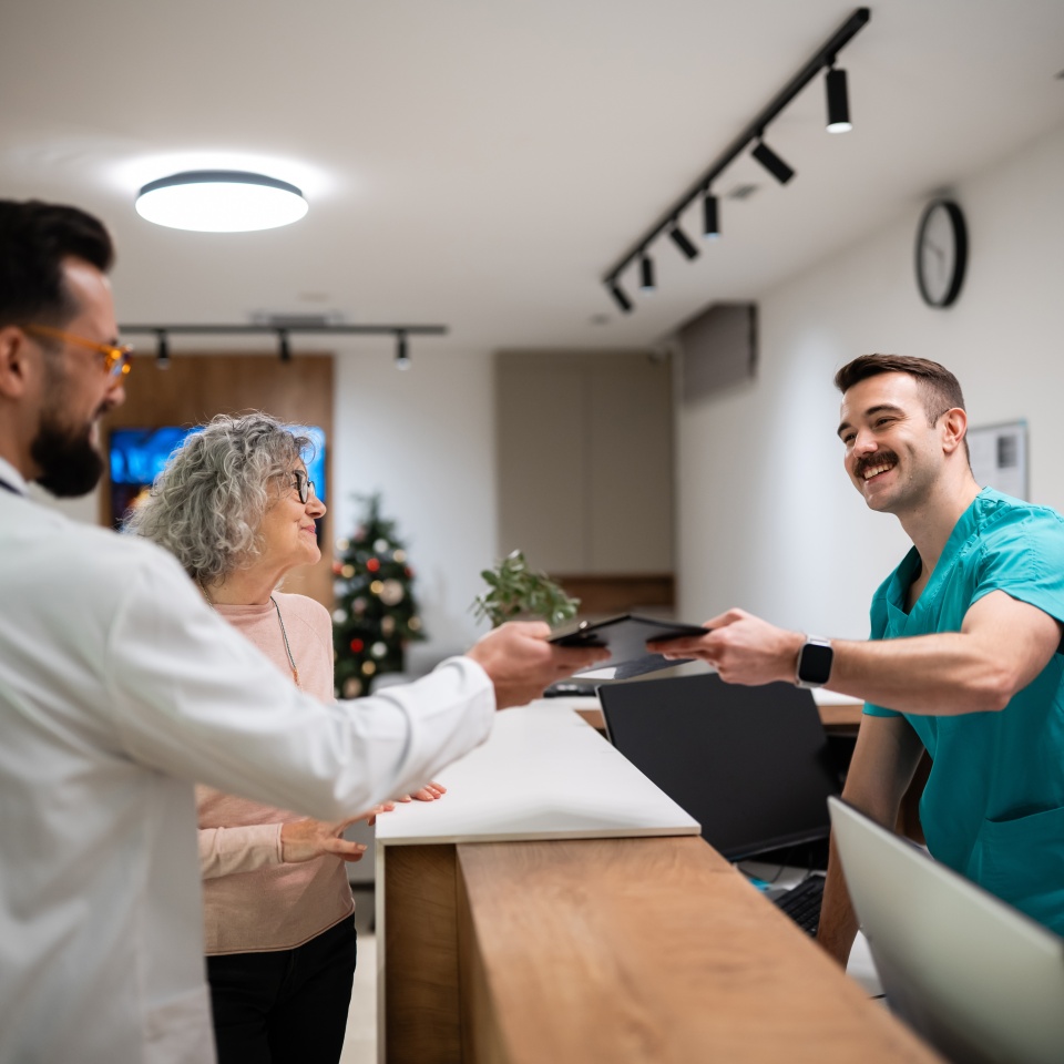 Doctor and nurse assisting a senior woman at a modern clinic reception, exchanging a digital tablet for patient registration
